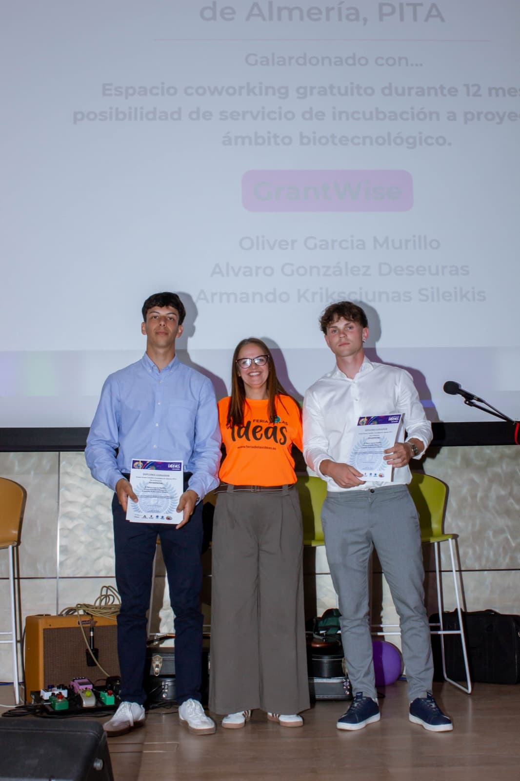 Armando y Óliver junto a la organización de la Feria de las Ideas durante la entrega del Premio PITA, con el detalle del galardón proyectado al fondo.